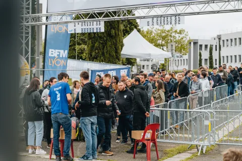 File d’attente à l’entrée du Festival, sous l’arche aux couleurs de BFM Lyon, encadrée par un bénévole en t-shirt Staff.
