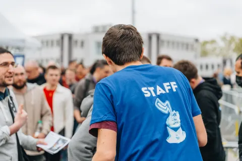 Bénévole de dos, t-shirt bleu « Staff » au logo du Festival, au milieu des visiteurs à l’accueil.