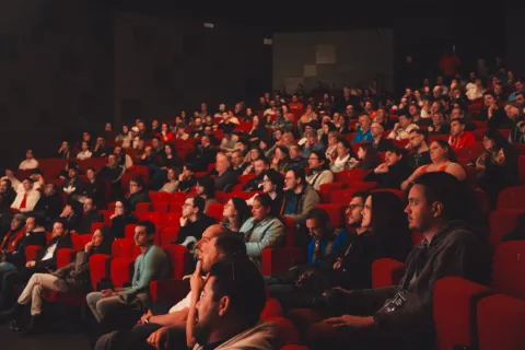 Salle de conférence comble, vue depuis le fond des gradins rouges.