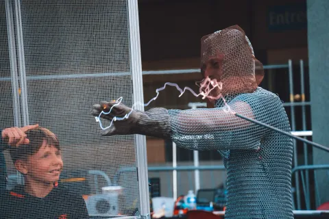 Démonstration de cage de Faraday : un homme en cotte de mailles touché par un arc électrique, un enfant fasciné à côté.