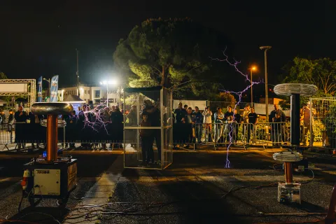 Bobines Tesla en action la nuit, des arcs violets devant le public derrière les barrières.