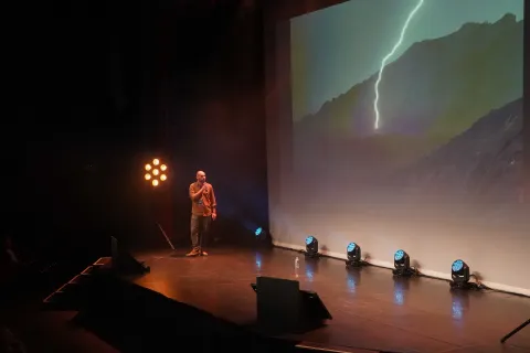 Intervenant seul sur scène devant la projection d’un éclair frappant une montagne.