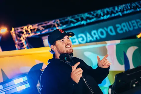 Animateur en casquette, micro et casque autour du cou, sur la scène extérieure aux couleurs du Festival, le soir.