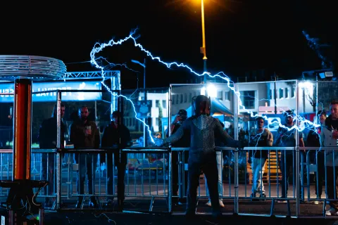 Bobines Tesla en action la nuit : un homme en combinaison métallique reçoit des arcs électriques devant le public.